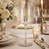 A clear wine glass with a small gray teddy bear figurine attached near the bowl, set on a decorated dining table with plates, cutlery, a candle, and flowers in the background.